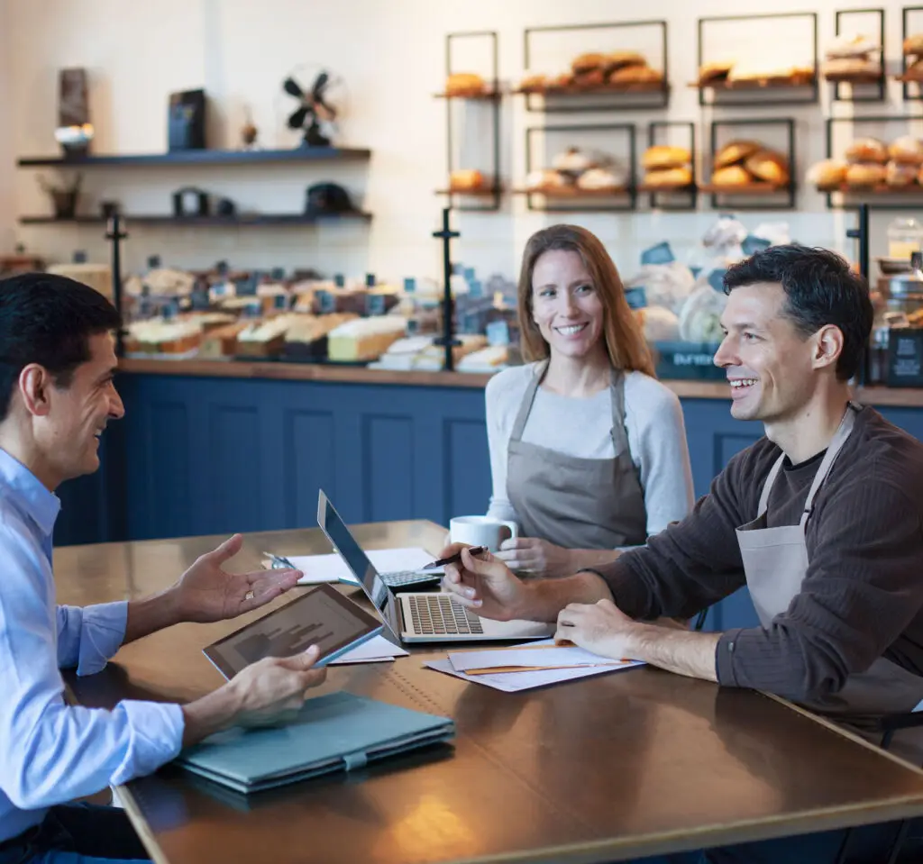 Mature Middle Eastern man financial advisor insurance agent talking advice with mature women and mature man owner staff employee sitting together in small business bakery cafe using laptop computer digital tablet planning