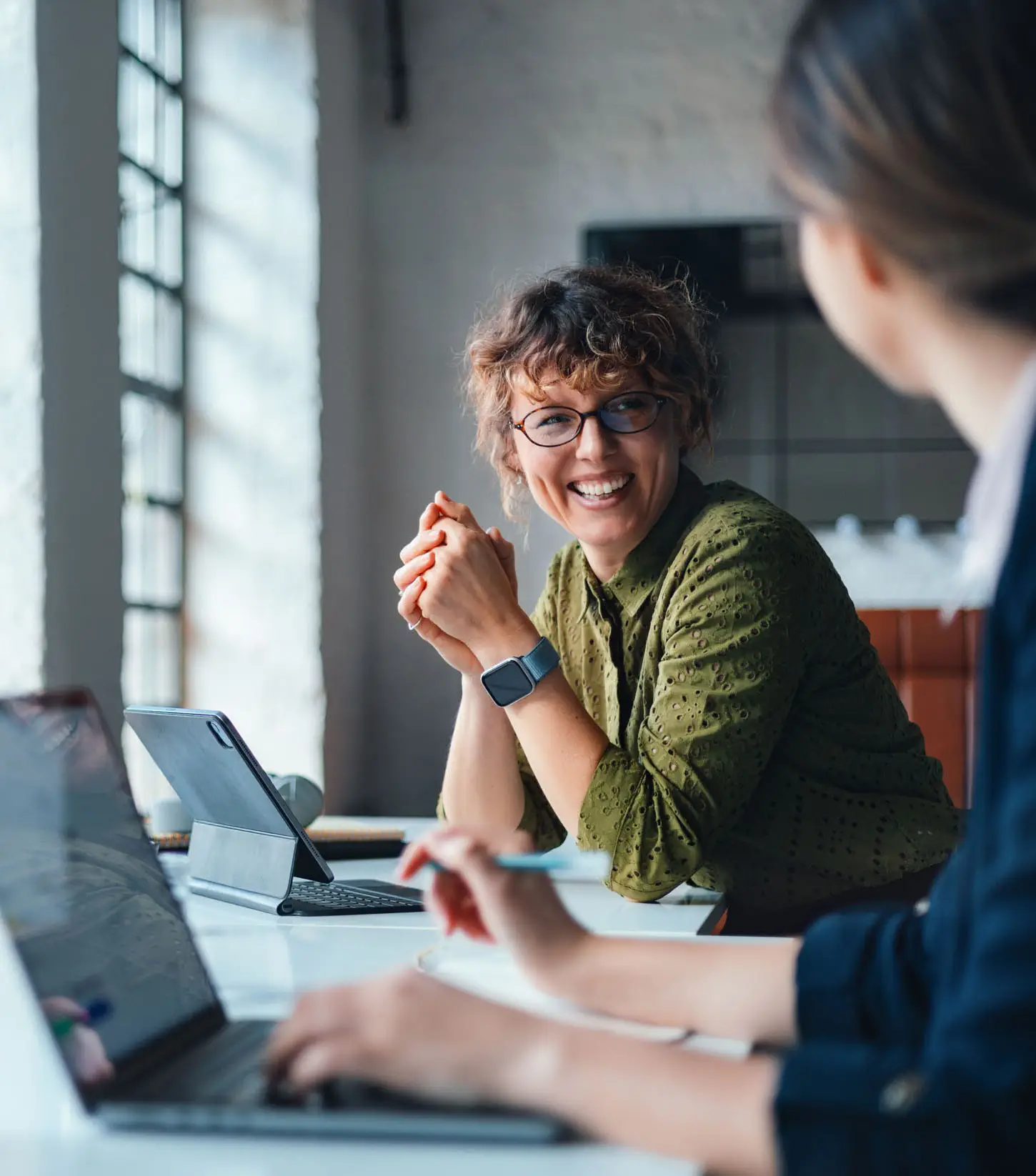 Two women collaborate in a bright coworking space, using laptops and a tablet. One smiles warmly, suggesting a positive, productive business discussion. Modern workspace, professional teamwork, and friendly corporate atmosphere.